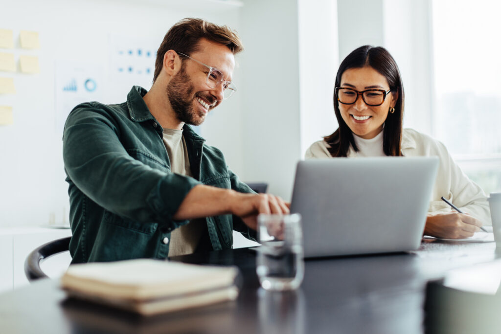 Two business people smiling and collaborating on credit union marketing strategies on a laptop in an office setting.