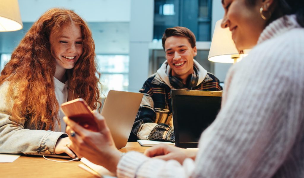 A group of Gen Z students studying together, with one using a smartphone, representing the need for accessible financial literacy tools from credit unions.